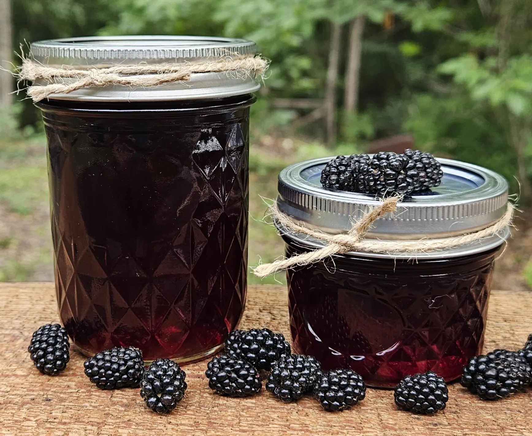 A glass jar of glossy blackberry jam on a wooden kitchen counter with fresh blackberries and a spoon beside it