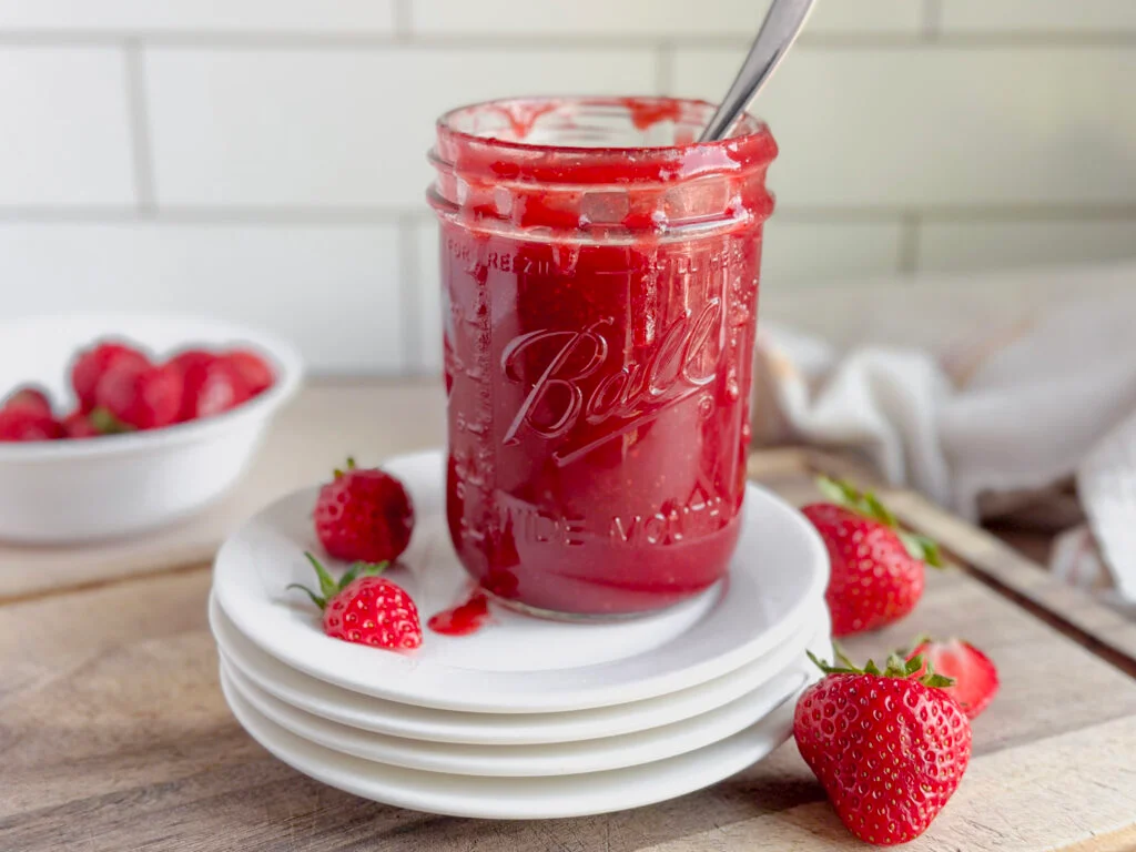 A glass jar of glossy homemade strawberry syrup on a light kitchen counter with fresh strawberries nearby, natural window light