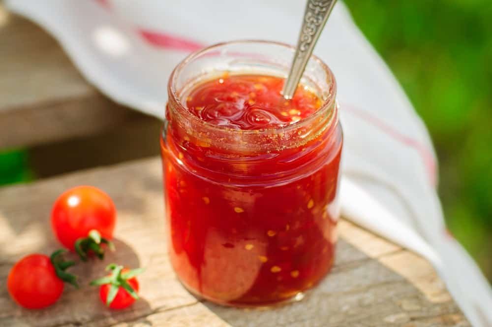 A glass jar of glossy tomato jam on a wooden kitchen counter with a spoon and slices of toasted bread nearby in warm natural window light