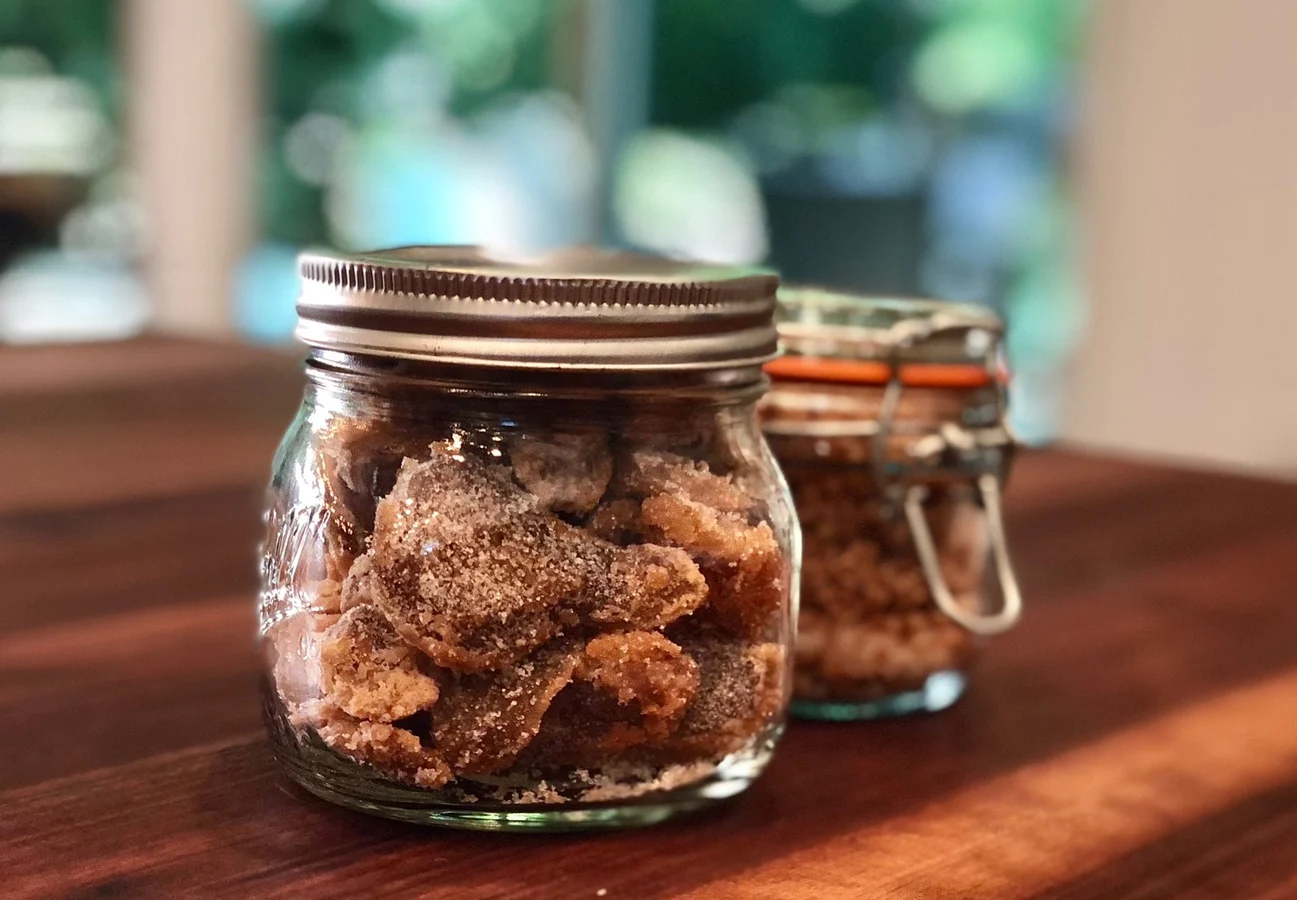 A glass jar of homemade candied ginger pieces with the lid off on a tidy kitchen counter, with a small spoon resting nearby