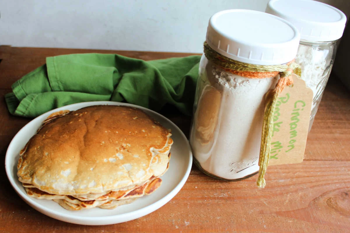 A glass jar of homemade pancake dry mix on a kitchen counter with a whisk, measuring cup, and a small bowl of sugar nearby