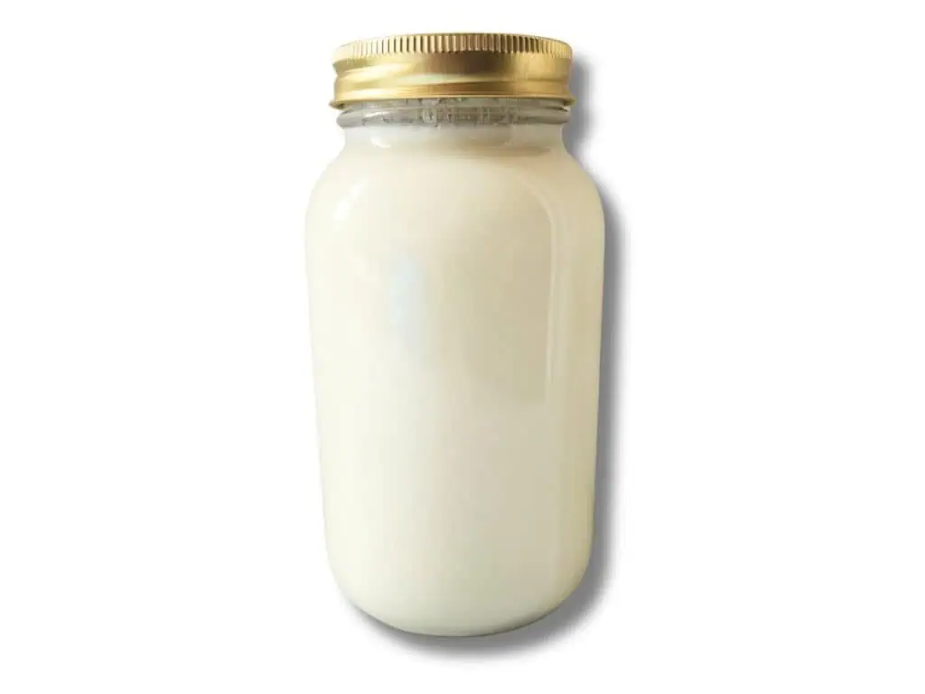A glass jar of thick, creamy cultured buttermilk on a bright kitchen counter beside a small whisk and a folded linen towel