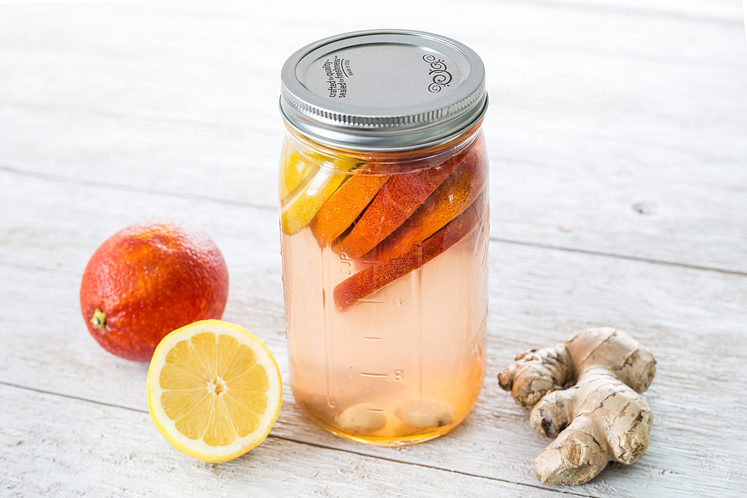 A glass mason jar filled with amber switchel over ice, with a lemon wheel and fresh ginger on a light kitchen counter in soft window light