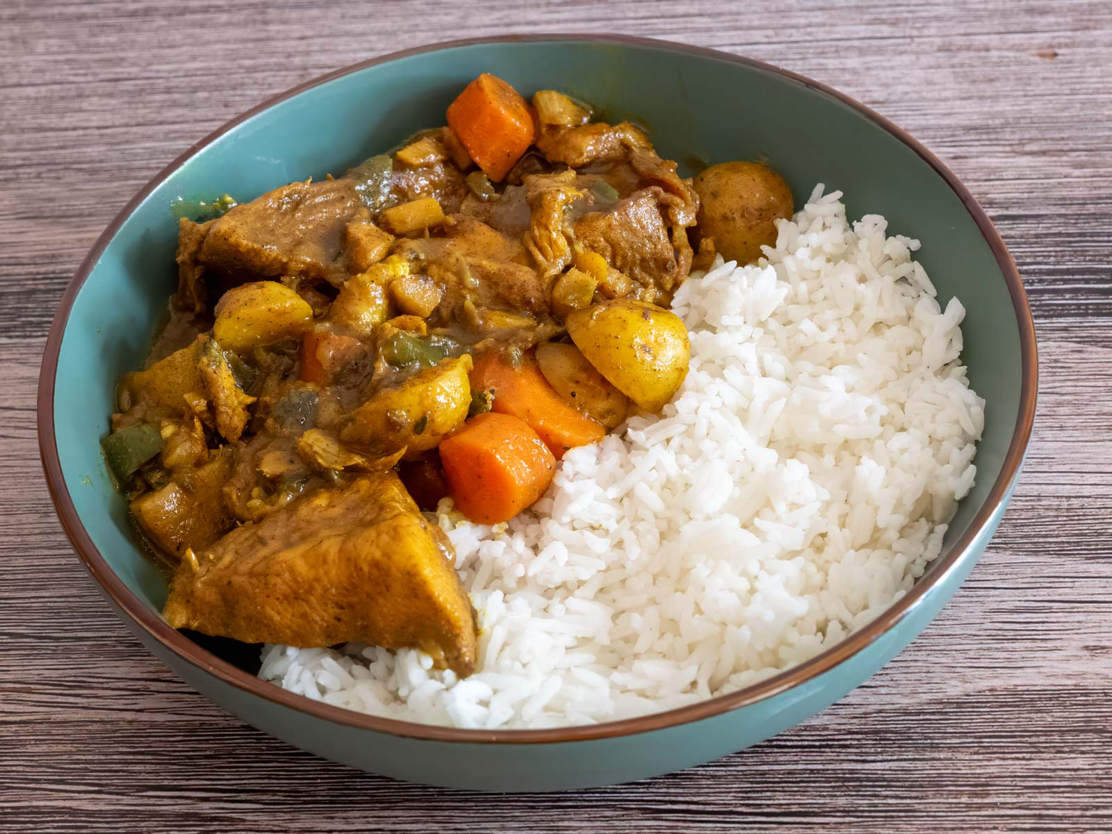 A glass meal prep container filled with Jamaican curry chicken and potatoes in golden sauce, resting on a kitchen counter with a lid nearby
