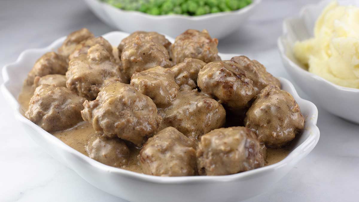 A glass meal prep container filled with Swedish meatballs and creamy gravy next to a small container of cooked egg noodles on a kitchen counter
