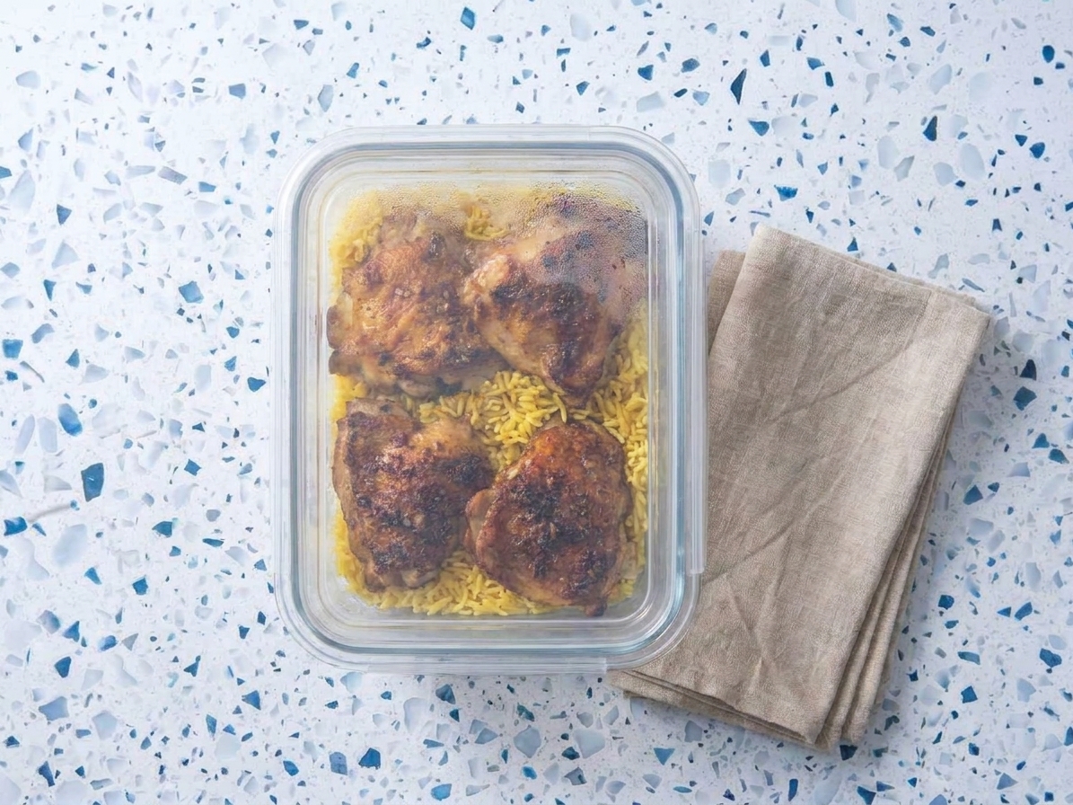 A glass meal prep container filled with chicken and yellow rice, cooled and ready to store with a tight-fitting lid on a kitchen counter
