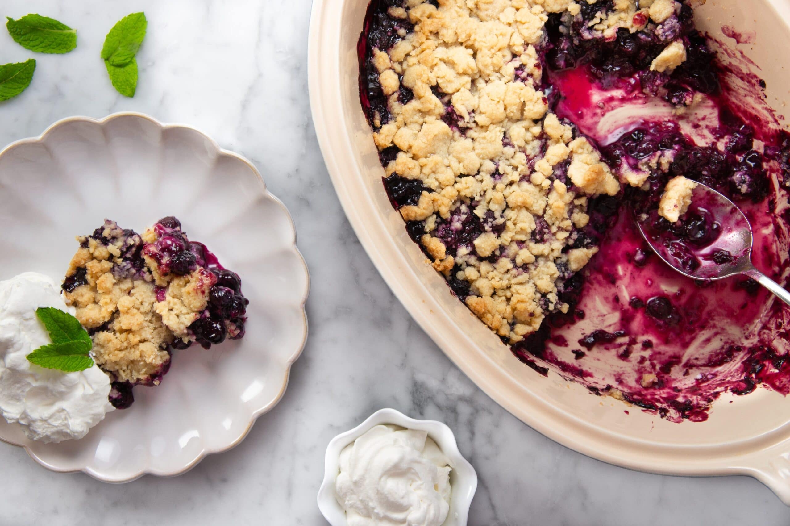 A glass meal prep container filled with leftover blueberry crumble, showing a layer of purple filling and crisp oat topping, sitting on a kitchen counter