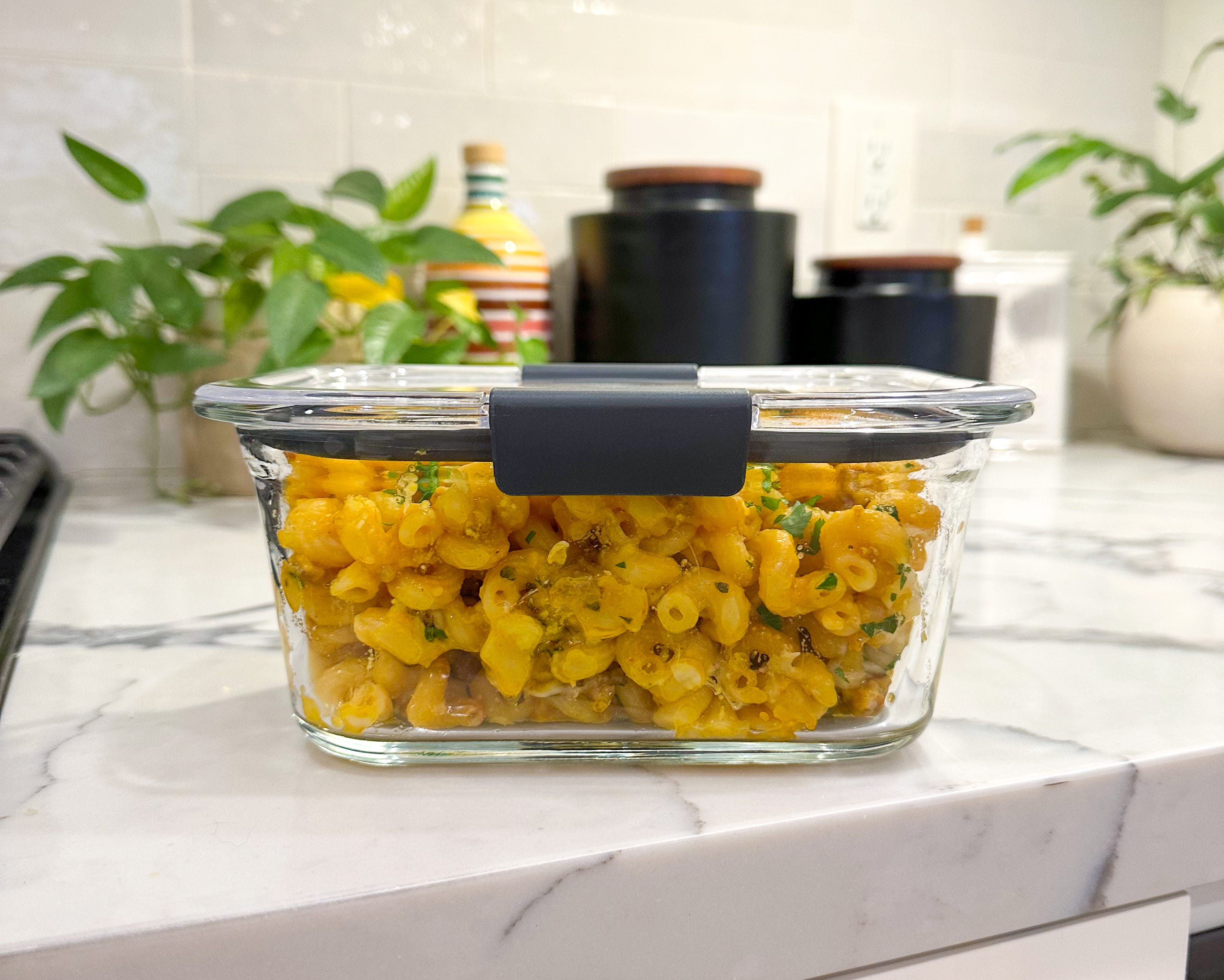 A glass meal prep container filled with leftover baked ziti topped with melted cheese, sitting on a kitchen counter with a lid beside it