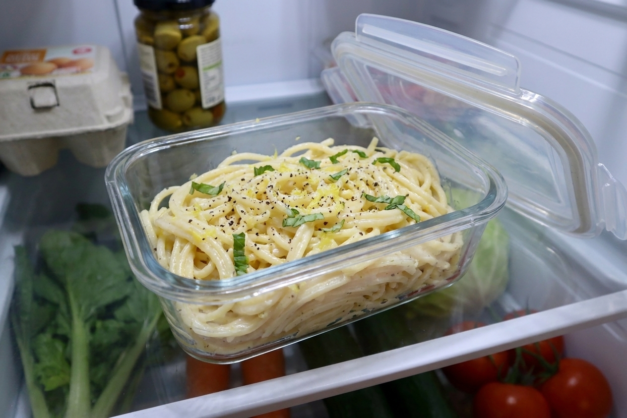 A glass meal prep container filled with leftover lemon garlic pasta on a refrigerator shelf, lid set beside it