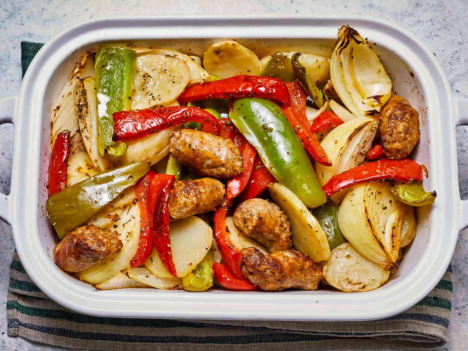 A glass meal prep container filled with leftover sausage, green peppers, onions, and tomato sauce with a lid set beside it on a kitchen counter