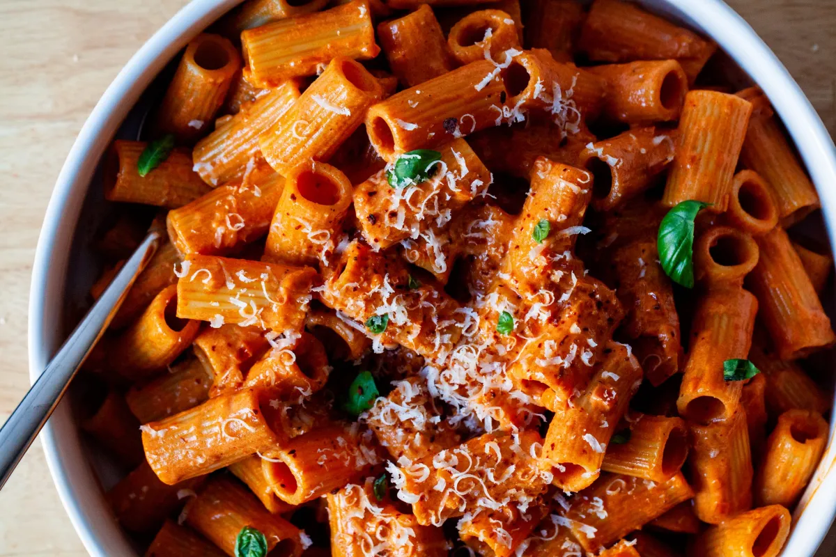 A glass meal prep container filled with leftover vodka rigatoni and sausage, lid set beside it on a kitchen counter