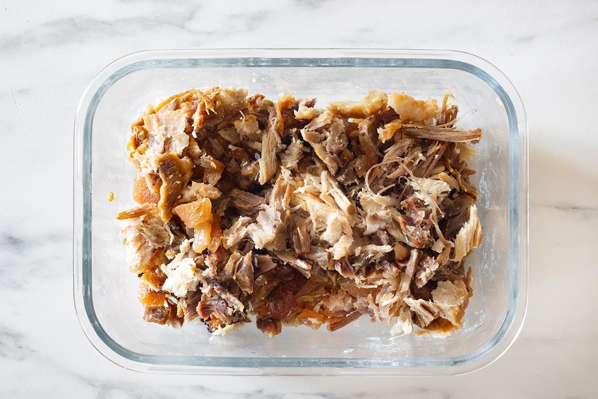 A glass meal prep container filled with shredded carnitas with a small container of cooking juices on a kitchen counter