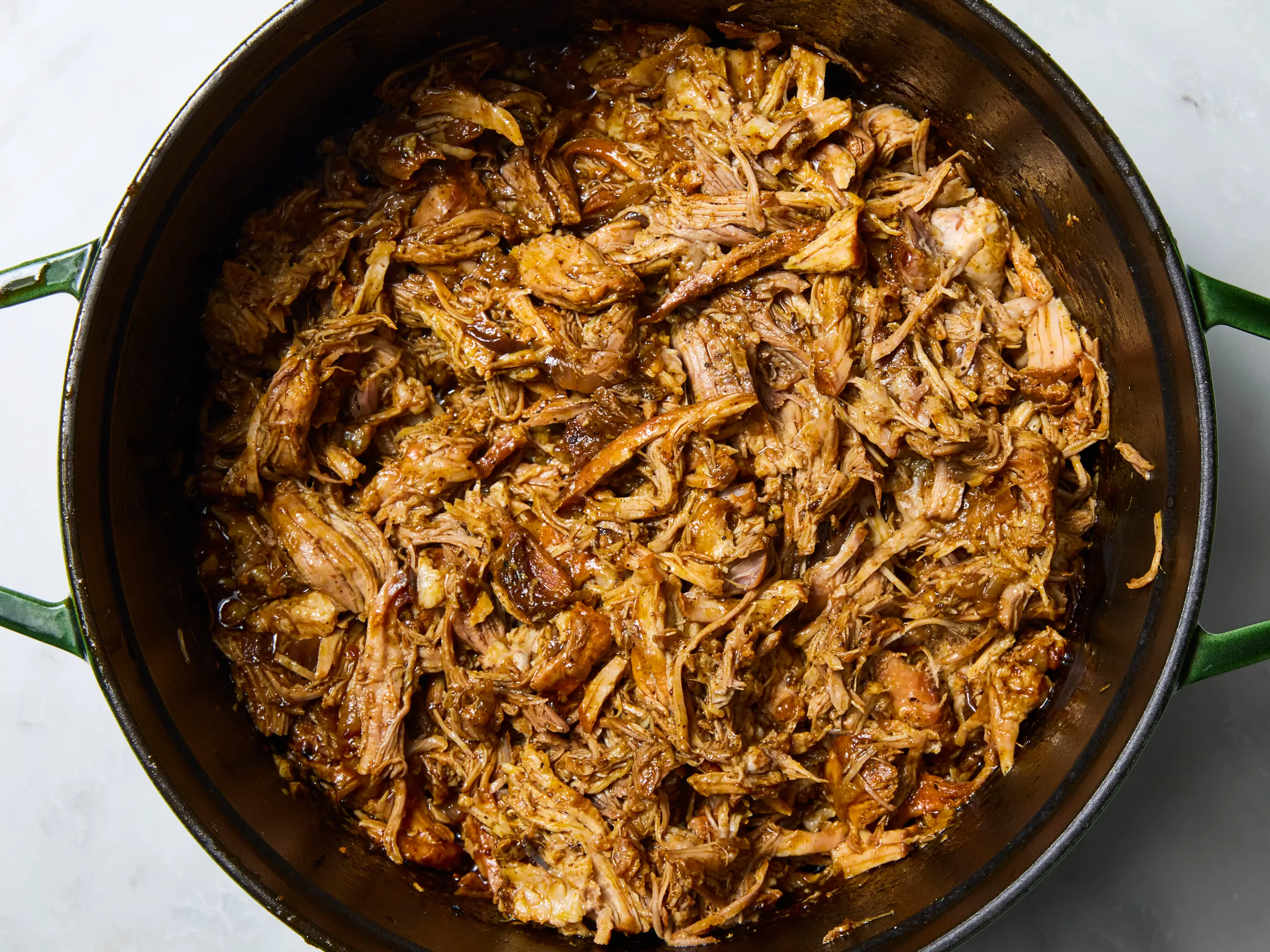 A glass meal prep container filled with shredded cochinita pibil and cooking juices, lid off on a kitchen counter with a spoon nearby