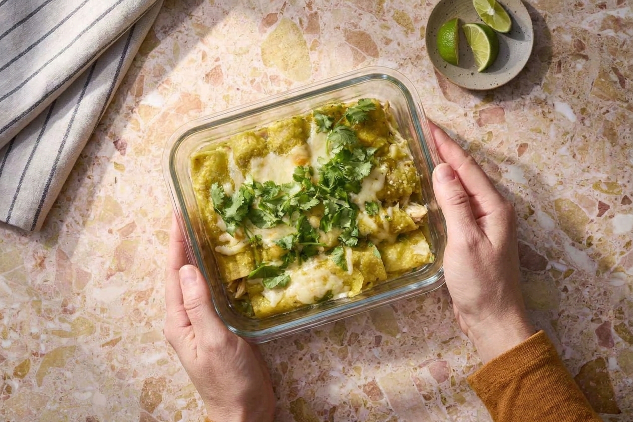 A glass meal prep container holding a portion of green chicken enchiladas with melted cheese and salsa verde on a kitchen counter