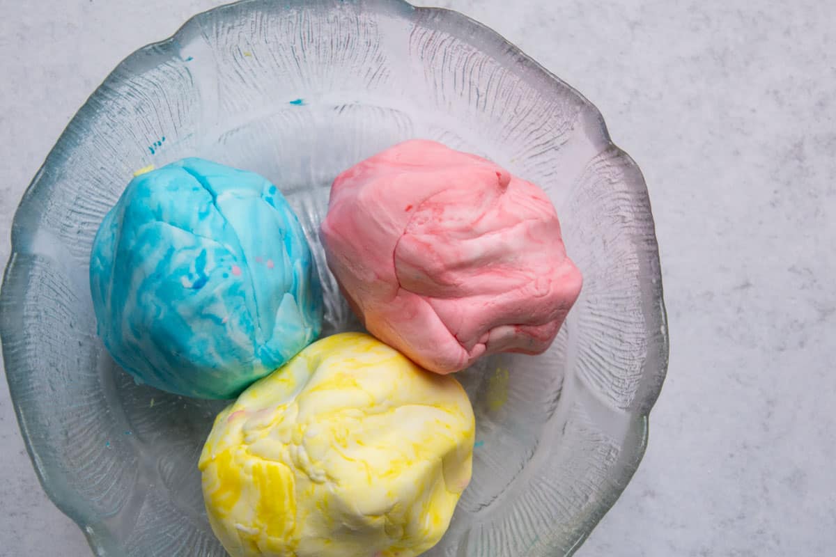 A glass mixing bowl filled with fluffy white cloud dough on a bright kitchen counter, with a wooden spoon and small bowls of flour and oil nearby