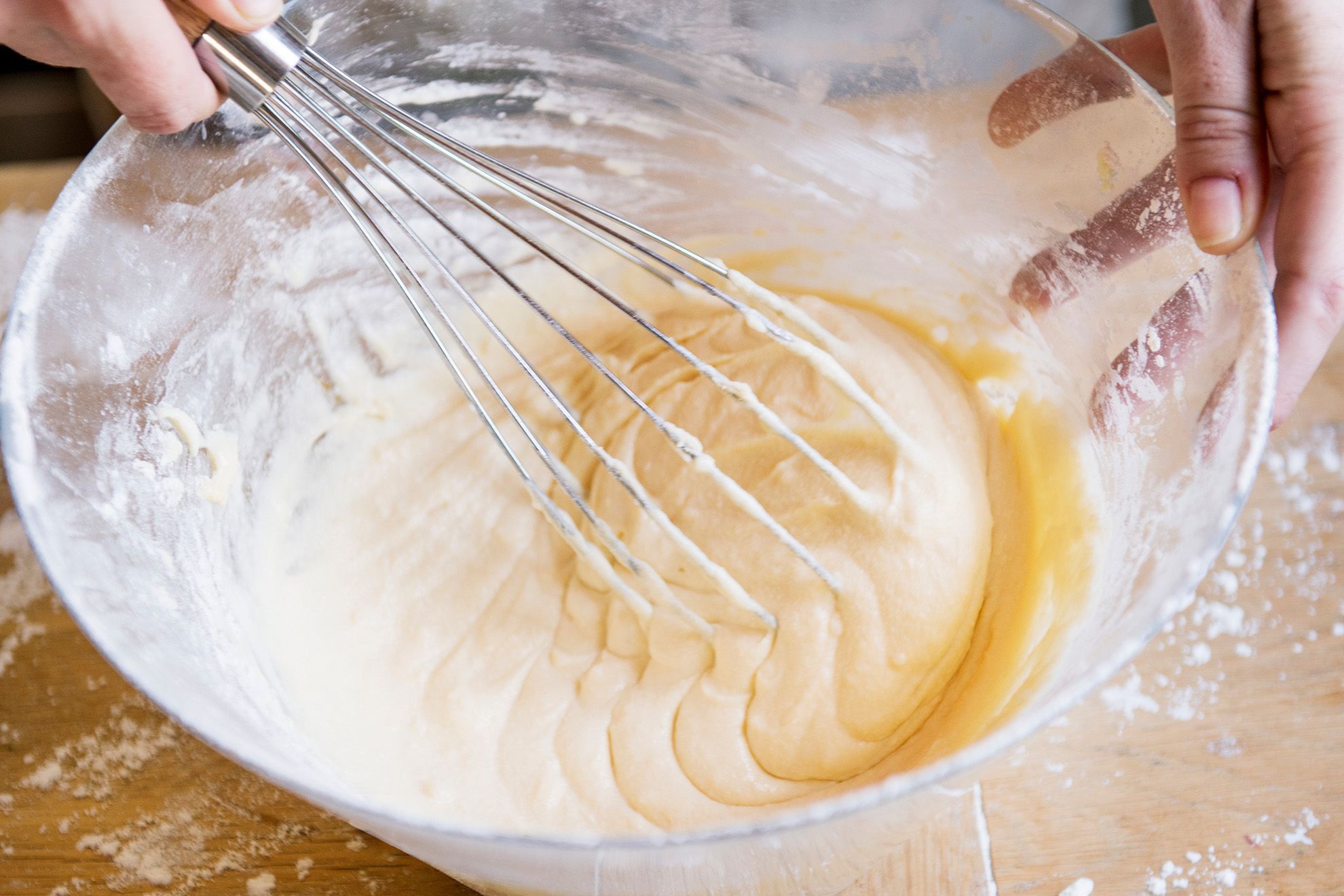 A glass mixing bowl filled with pineapple cake batter being whisked on a kitchen counter with measuring spoons nearby