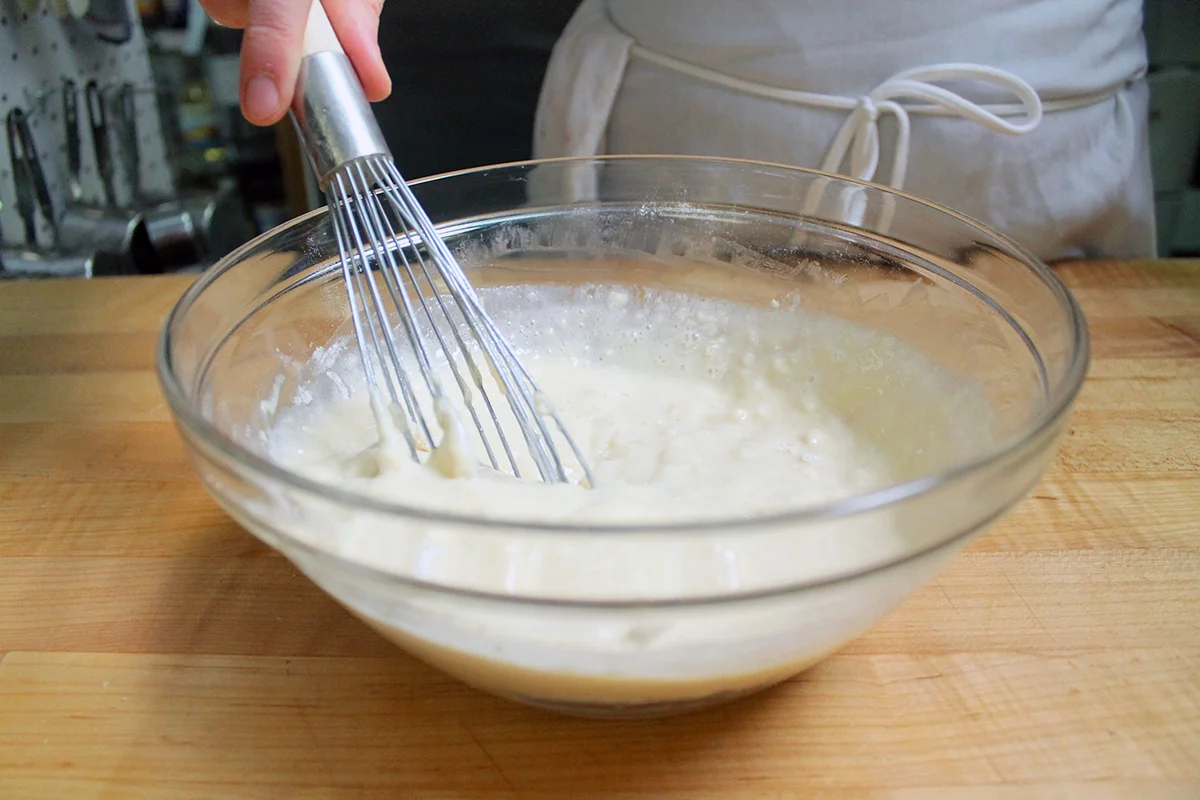 A glass mixing bowl of pancake batter being stirred on a clean kitchen counter with flour, sugar, and baking soda nearby