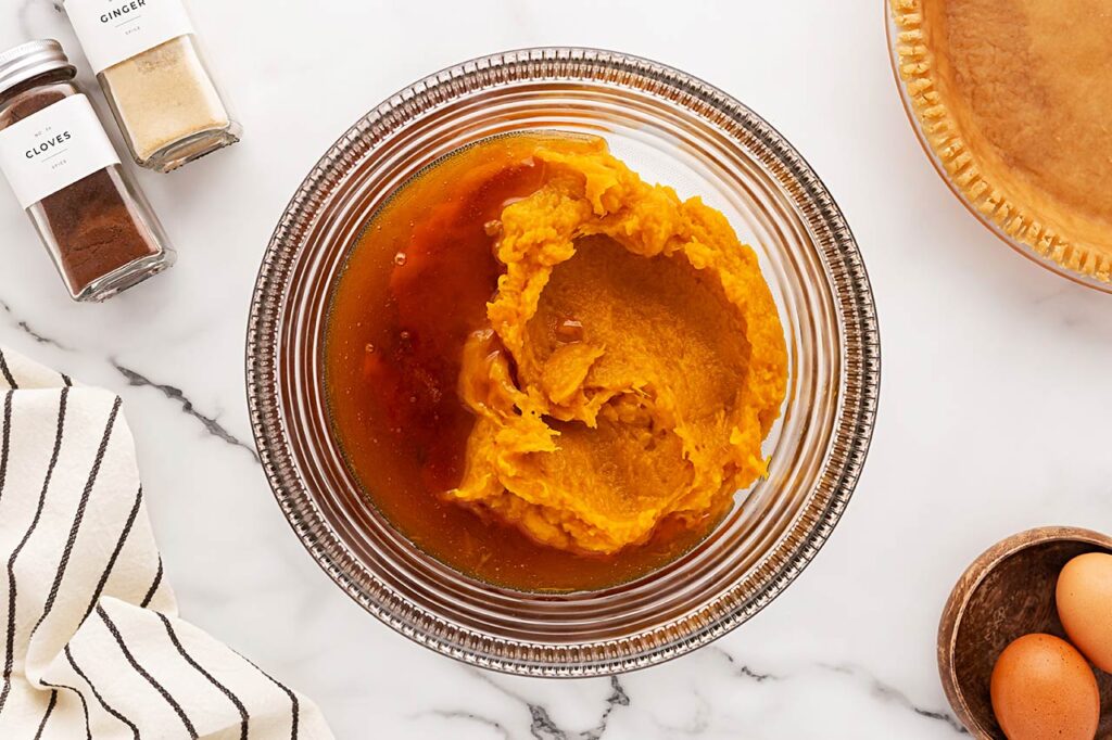 A glass mixing bowl of smooth pumpkin pie filling with visible cinnamon and nutmeg on top on a warm-toned kitchen counter