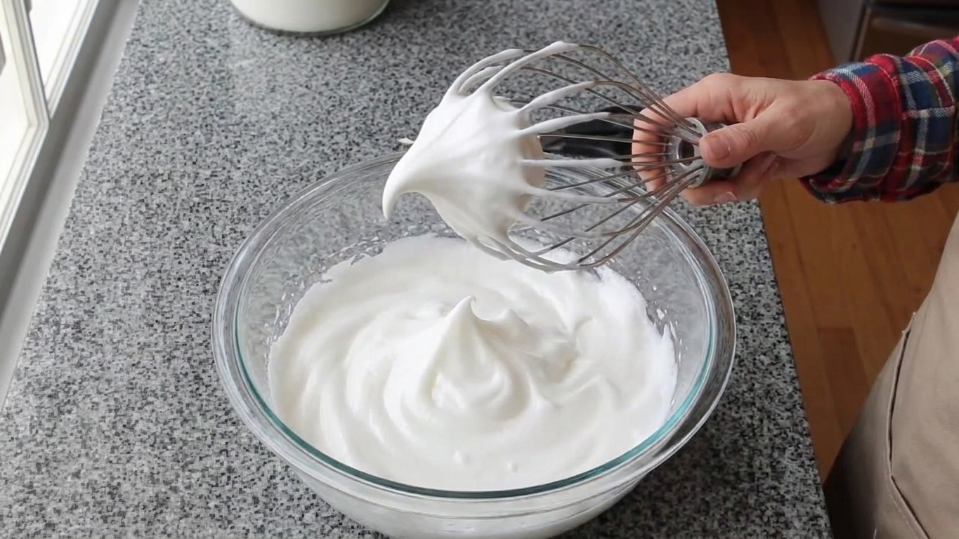 A glass mixing bowl of whipped egg whites holding glossy medium-stiff peaks on a whisk