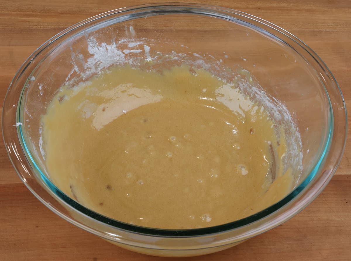 A glass mixing bowl with blondie batter being stirred with a rubber spatula on a kitchen counter, chocolate chips nearby, warm indoor lighting