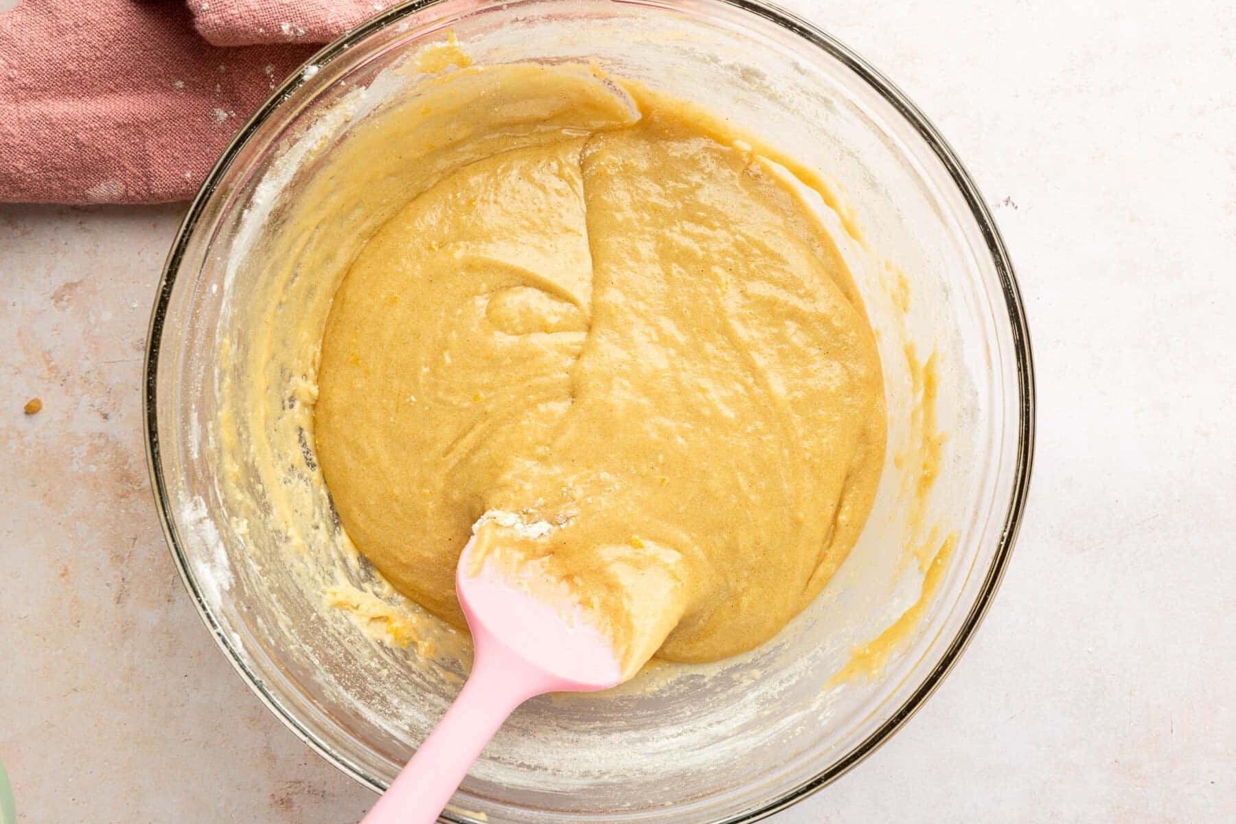 A glass mixing bowl with glossy blondie batter being stirred with a spatula on a kitchen counter