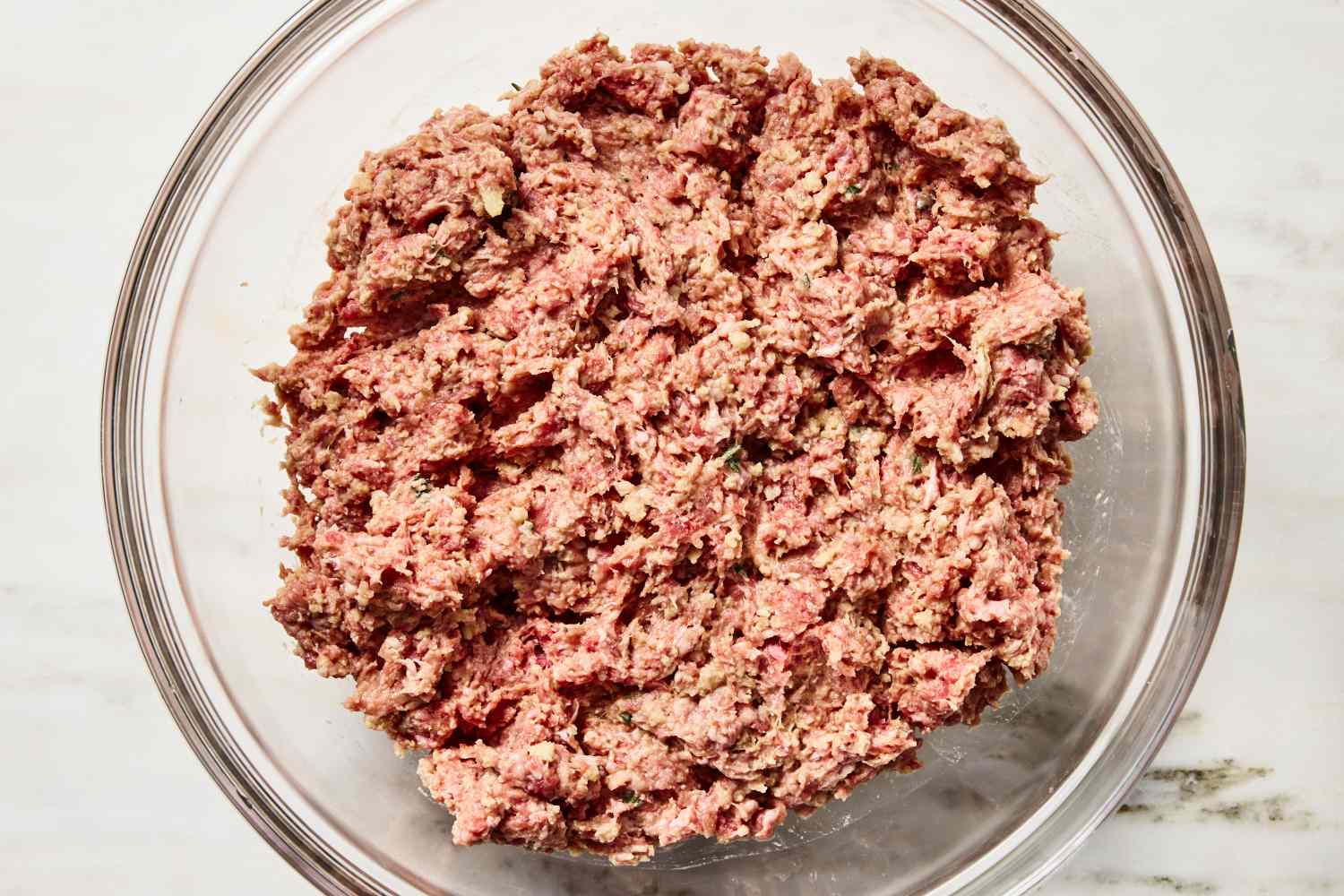 A glass mixing bowl with ground beef, breadcrumbs, egg, grated onion, and seasonings partially mixed on a kitchen counter