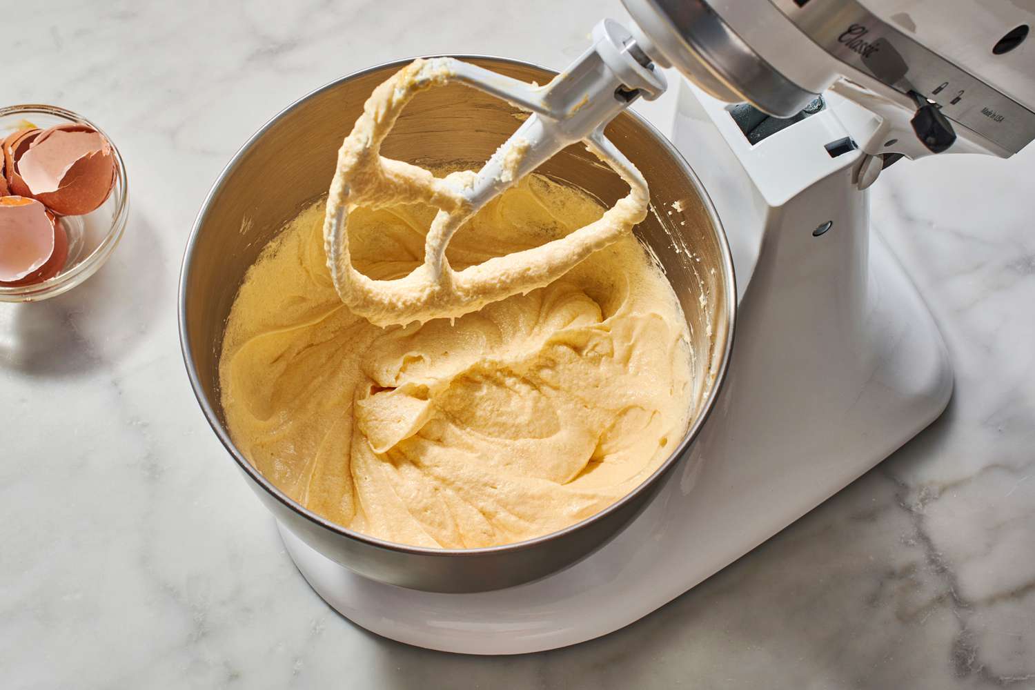 A glass mixing bowl with pale yellow cake batter being whisked until smooth on a bright kitchen counter