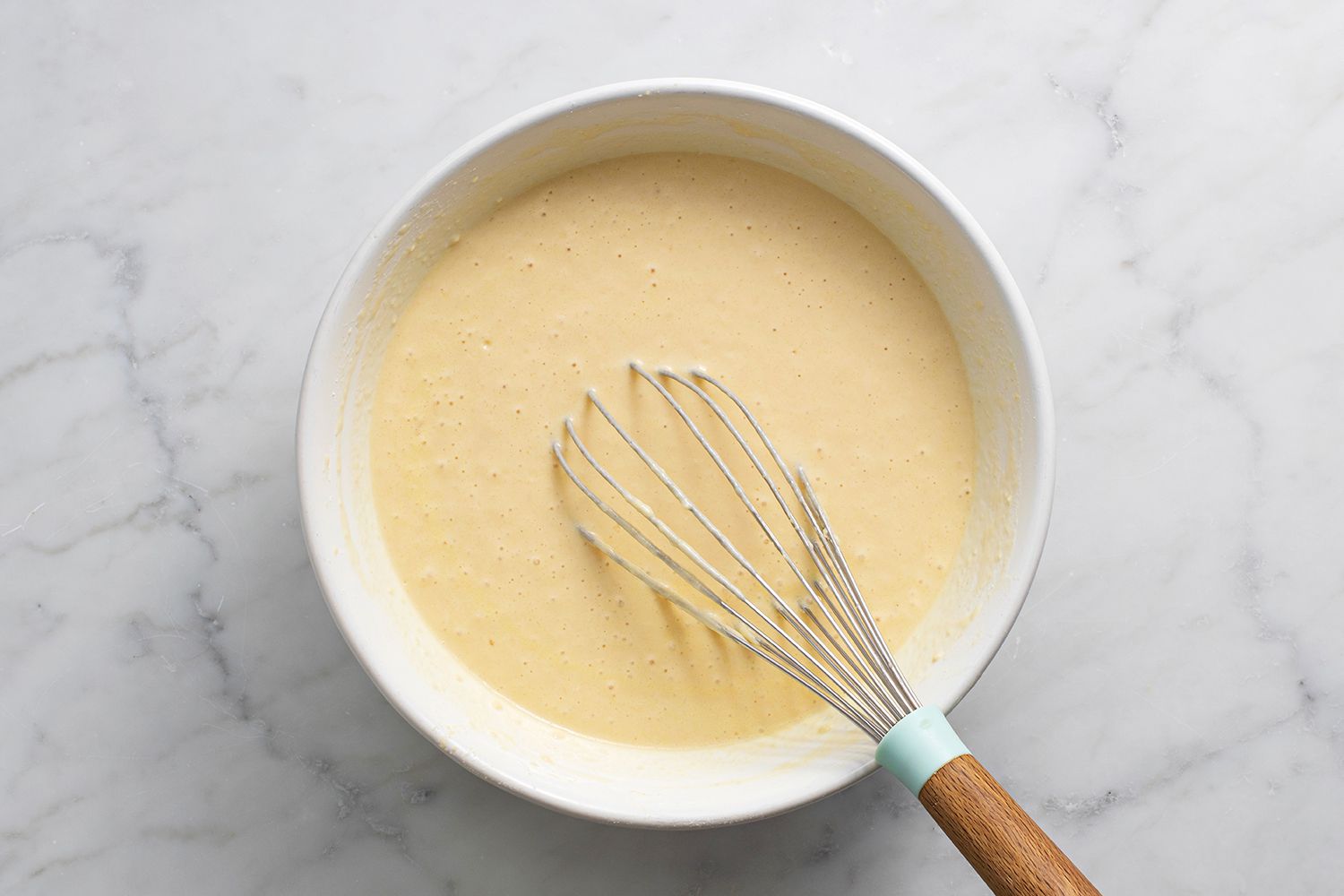 A glass mixing bowl with waffle batter being whisked until just combined on a tidy kitchen counter