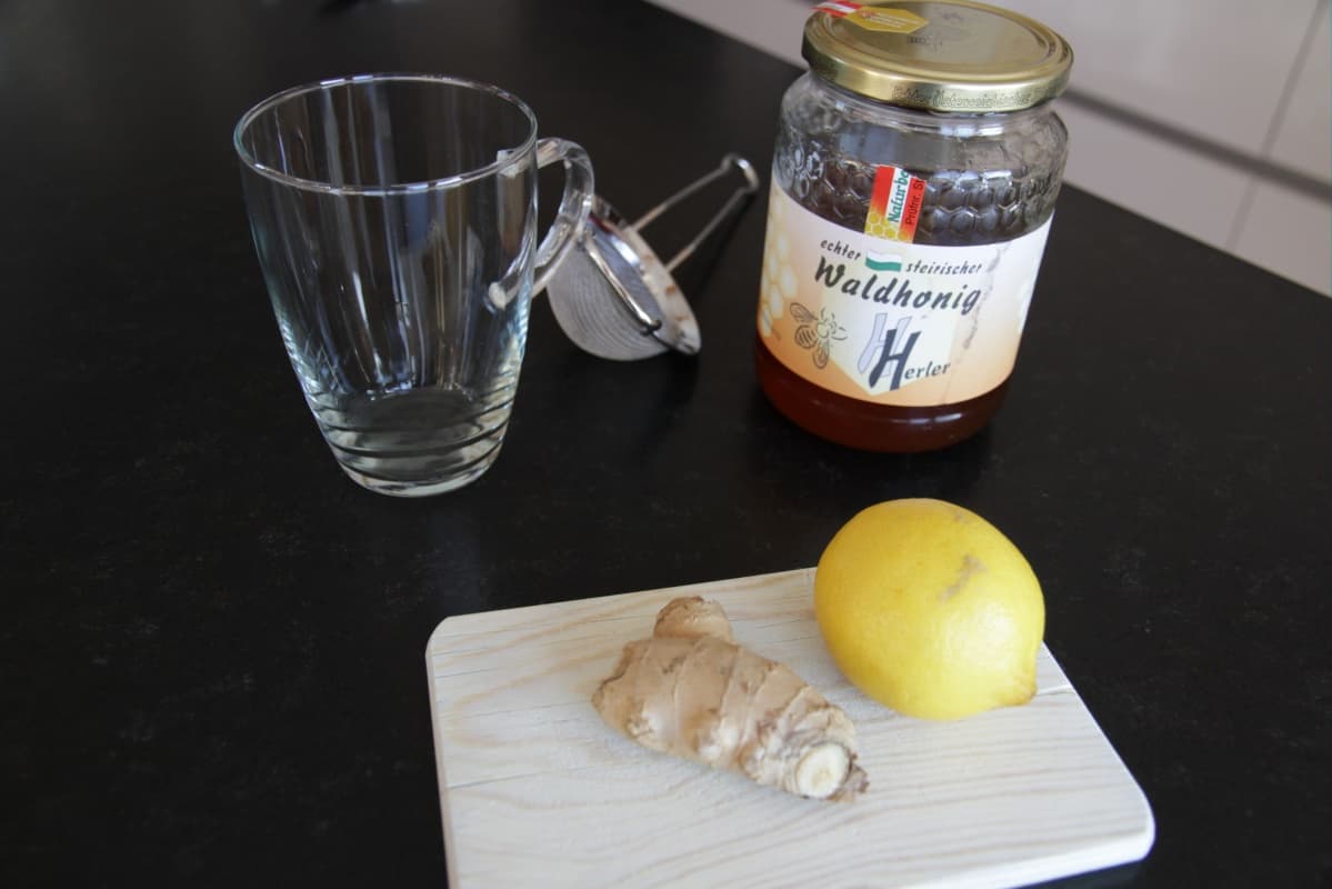 A glass mug of warm ginger water with lemon slices on a light stone kitchen counter, with a small bowl of sliced fresh ginger and a honey jar nearby