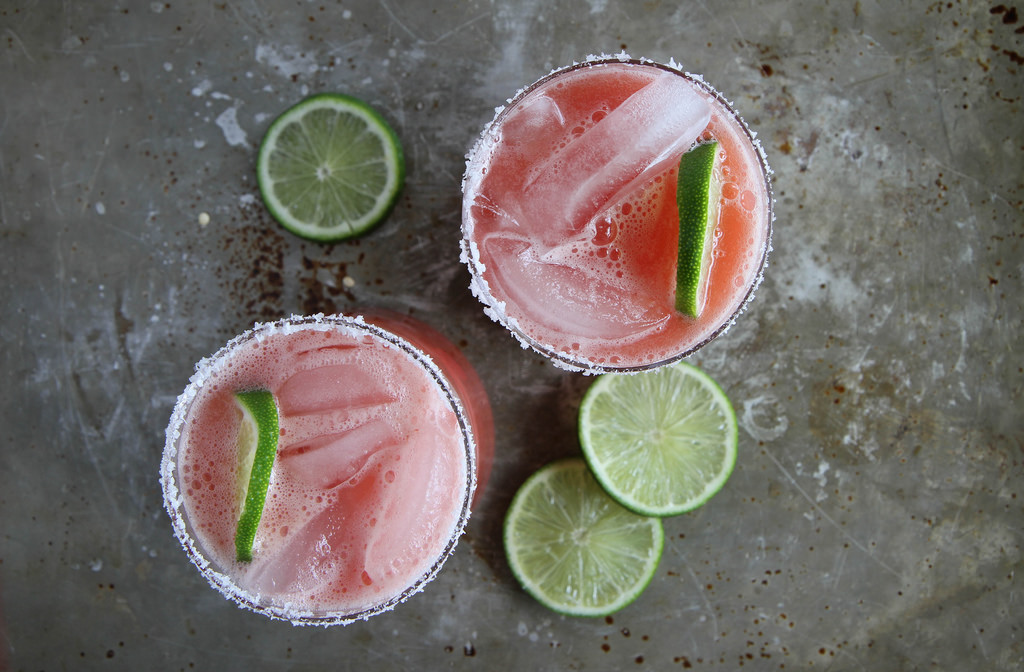 A glass of watermelon margarita on the rocks with a salted rim, lime wedge, and small watermelon slice on a bright kitchen counter
