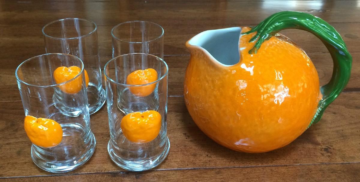 A glass pitcher of orange juice on a kitchen counter next to empty highball glasses and a bowl of ice, ready for serving