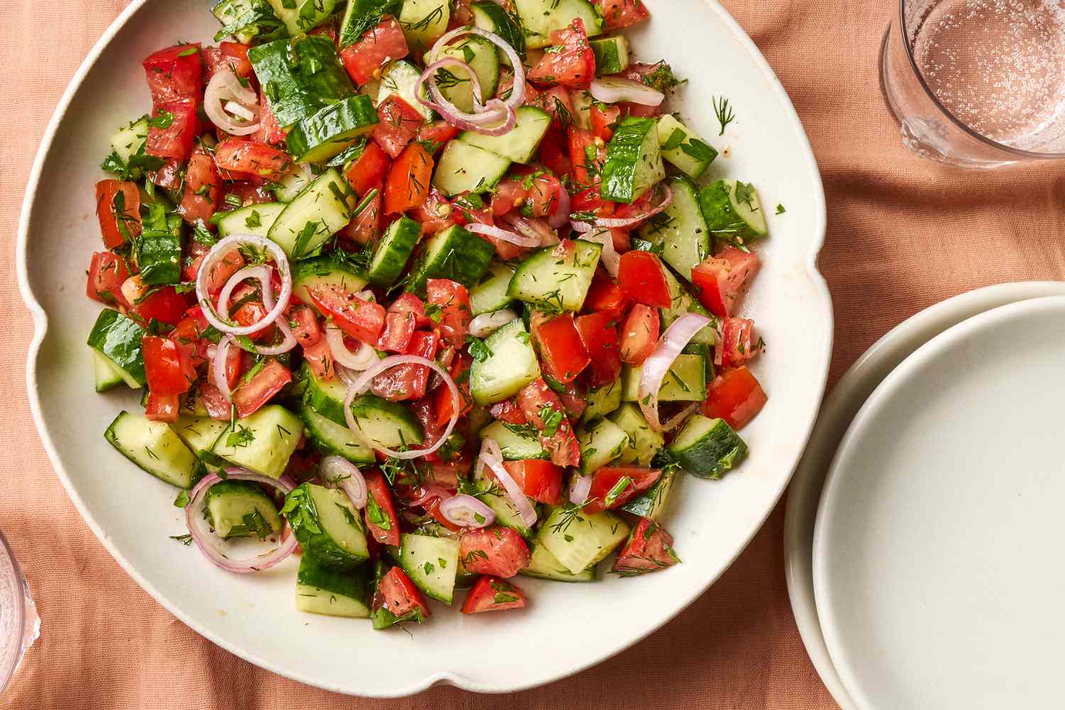 A glass serving bowl filled with tomato cucumber salad with red onion and fresh dill on a light stone kitchen counter, natural window light