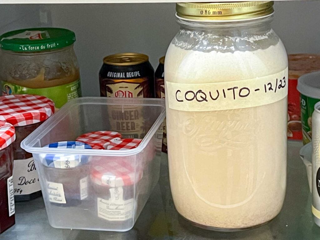 A glass swing-top bottle filled with coquito sitting on a refrigerator shelf next to cinnamon sticks and a small jar of nutmeg