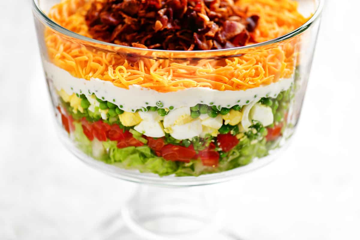 A glass trifle dish filled with a classic-style seven layer salad showing distinct layers of iceberg lettuce, green peas, diced celery, red bell pepper, creamy dressing, crisp bacon, and shredded cheddar cheese, natural window light, real food photography
