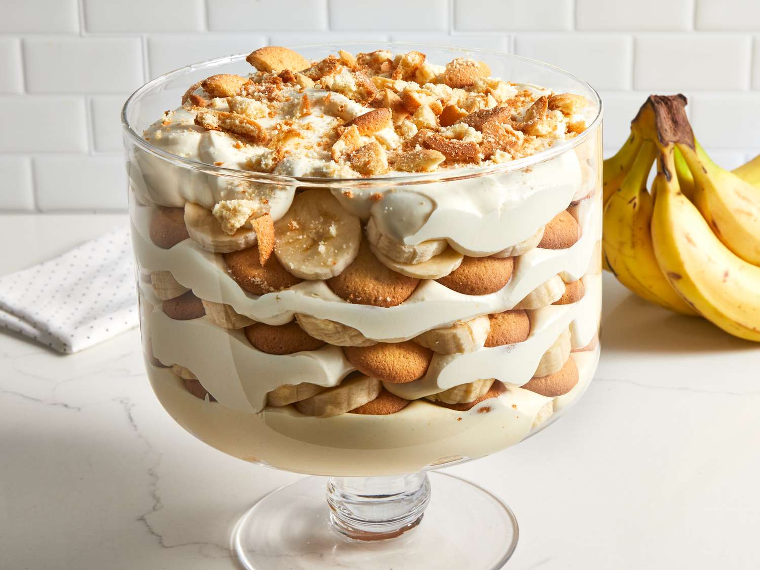 A glass trifle dish filled with layered banana pudding, sliced bananas, and Nilla Wafers, topped with whipped cream on a bright kitchen counter