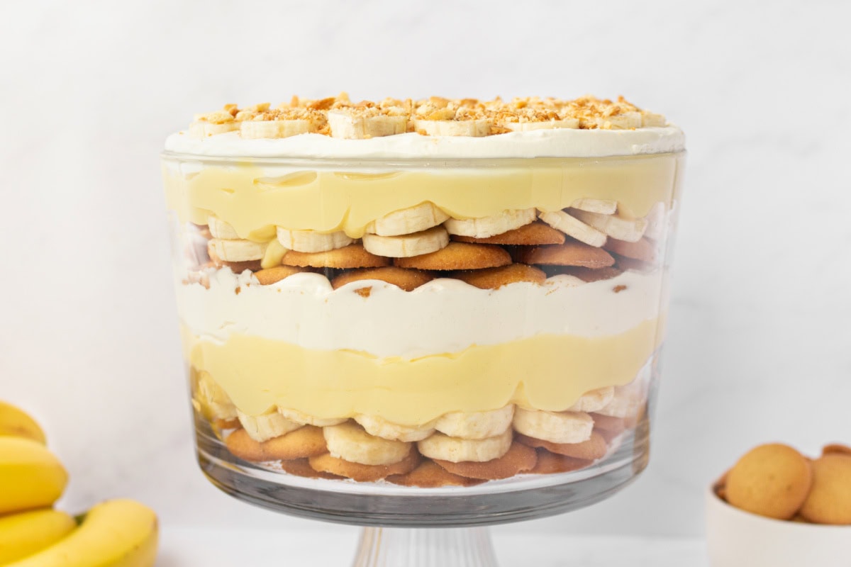 A glass trifle dish filled with layered banana pudding, sliced bananas, and vanilla wafers on a bright kitchen counter