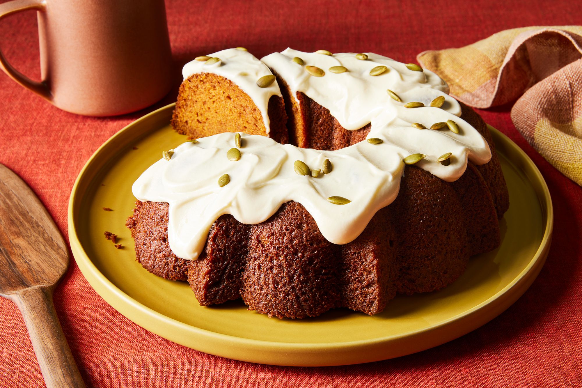 A glazed pumpkin bundt cake on a ceramic cake stand on a bright kitchen counter, with a few slices cut to show the moist orange crumb