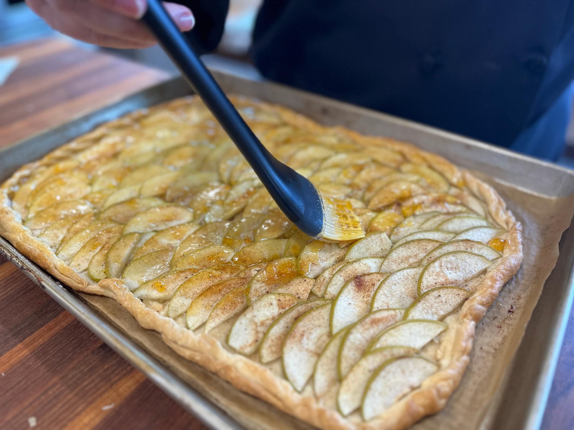 A glossy French-style apple tart with thinly fanned apple slices on a buttery crust, set on parchment on a baking sheet in a warm kitchen