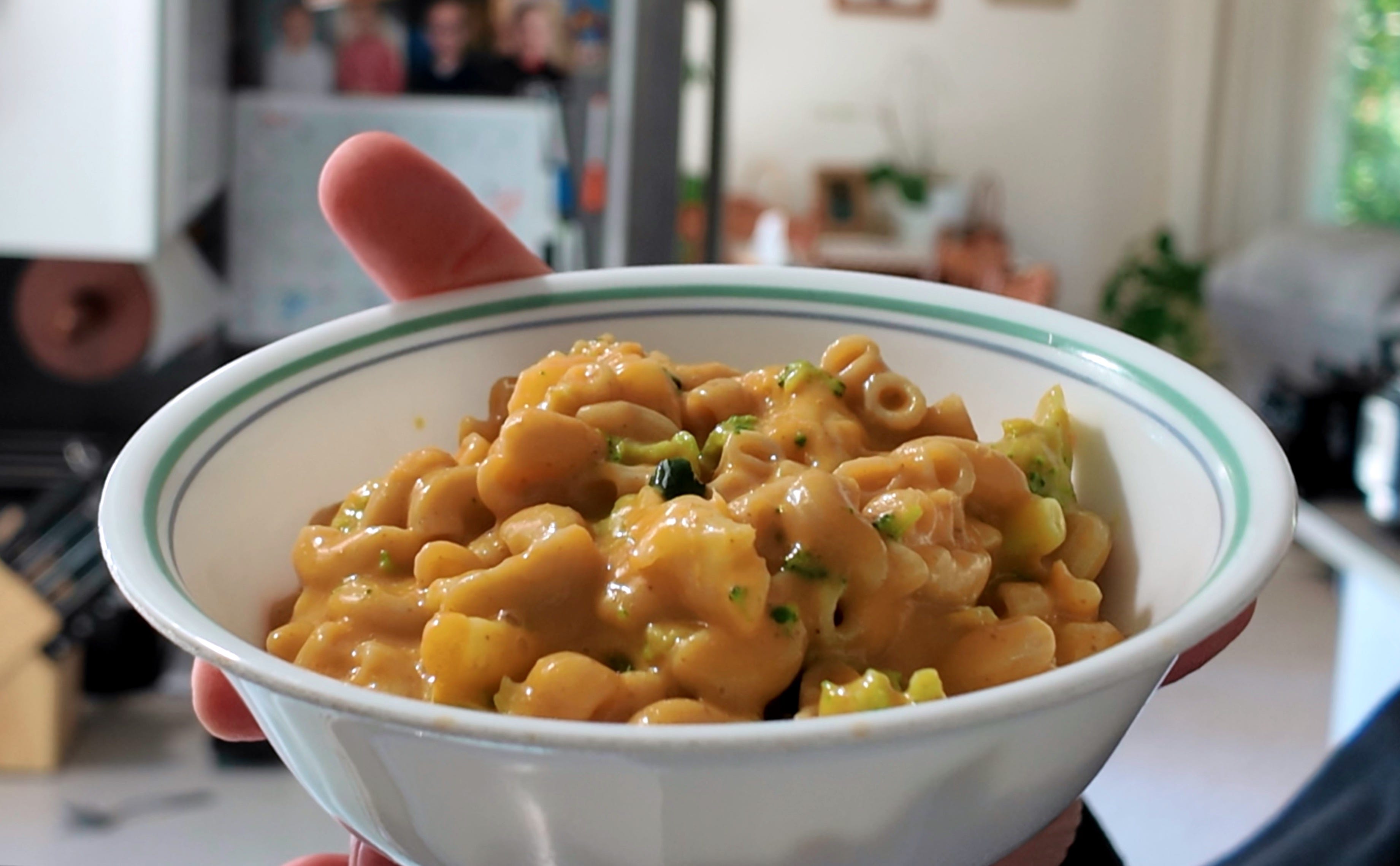 A glossy bowl of creamy vegan mac and cheese with a golden, lightly toasted breadcrumb topping on a wooden kitchen table in soft natural light