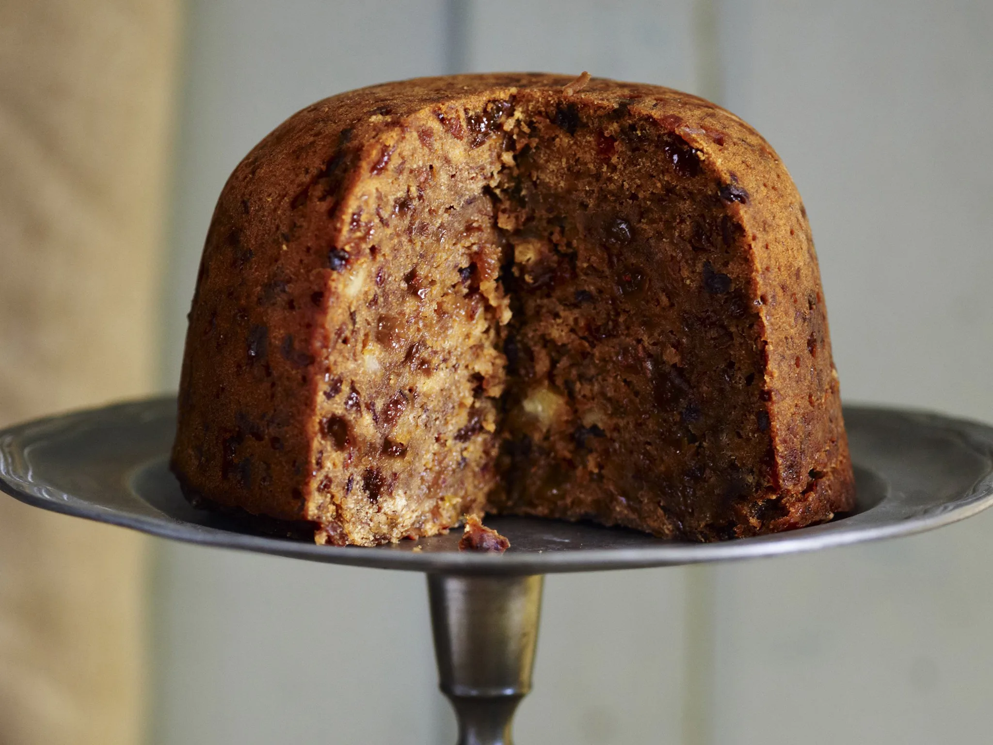 A glossy, dark steamed plum pudding unmolded onto a small plate with warm brandy sauce spooned over the top, on a wooden table with soft kitchen lighting
