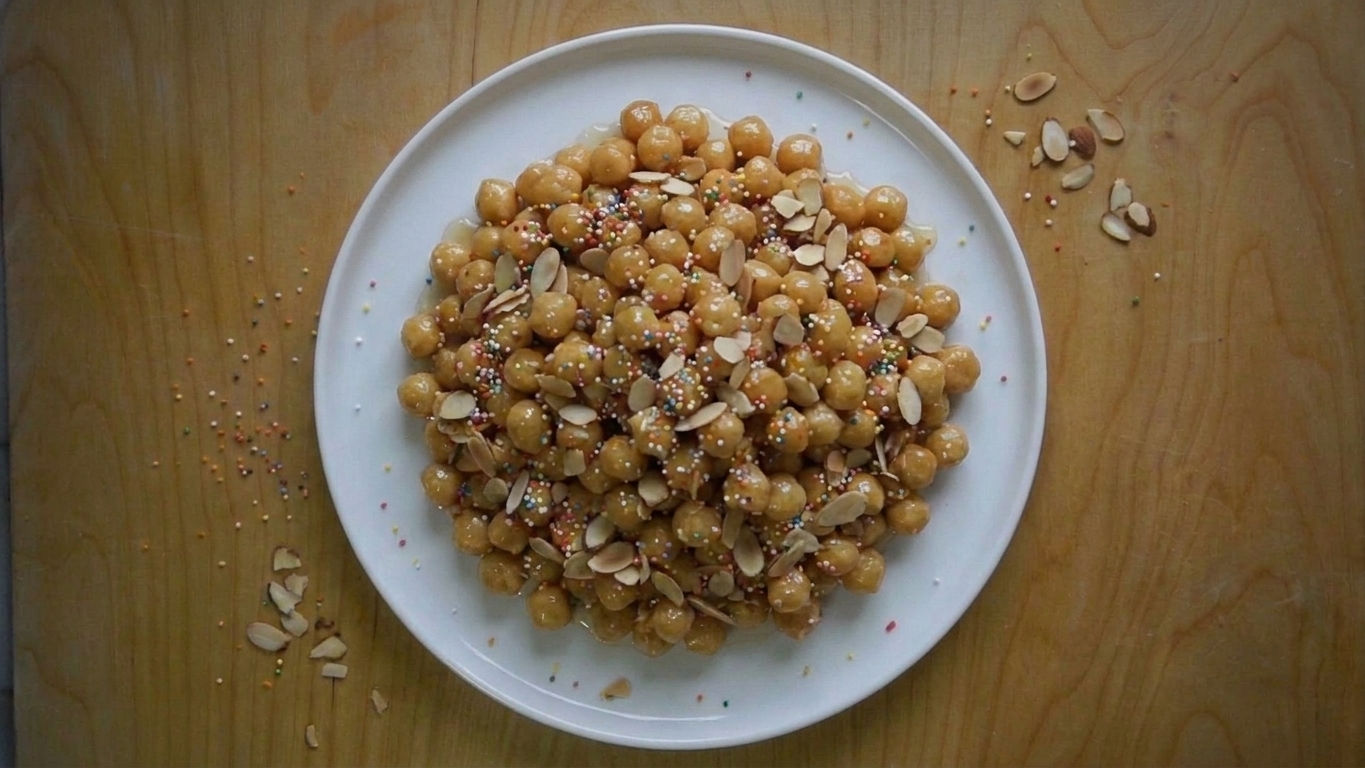A glossy mound of Italian struffoli (tiny honey-glazed fried dough balls) piled on a white platter with colorful sprinkles and toasted sliced almonds on a warm kitchen table