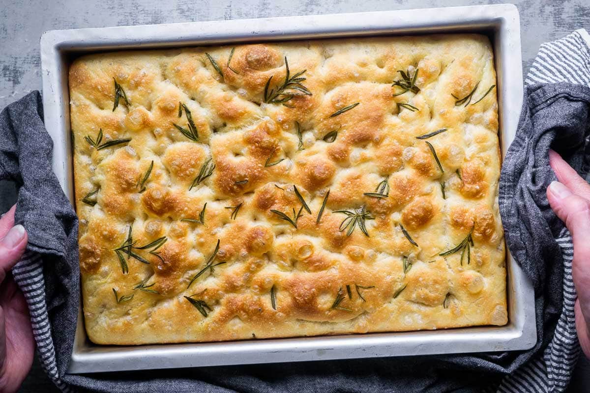 A golden Italian rosemary focaccia baked in a metal pan with a glossy olive oil top, deep dimples, and flaky salt on a warm kitchen counter