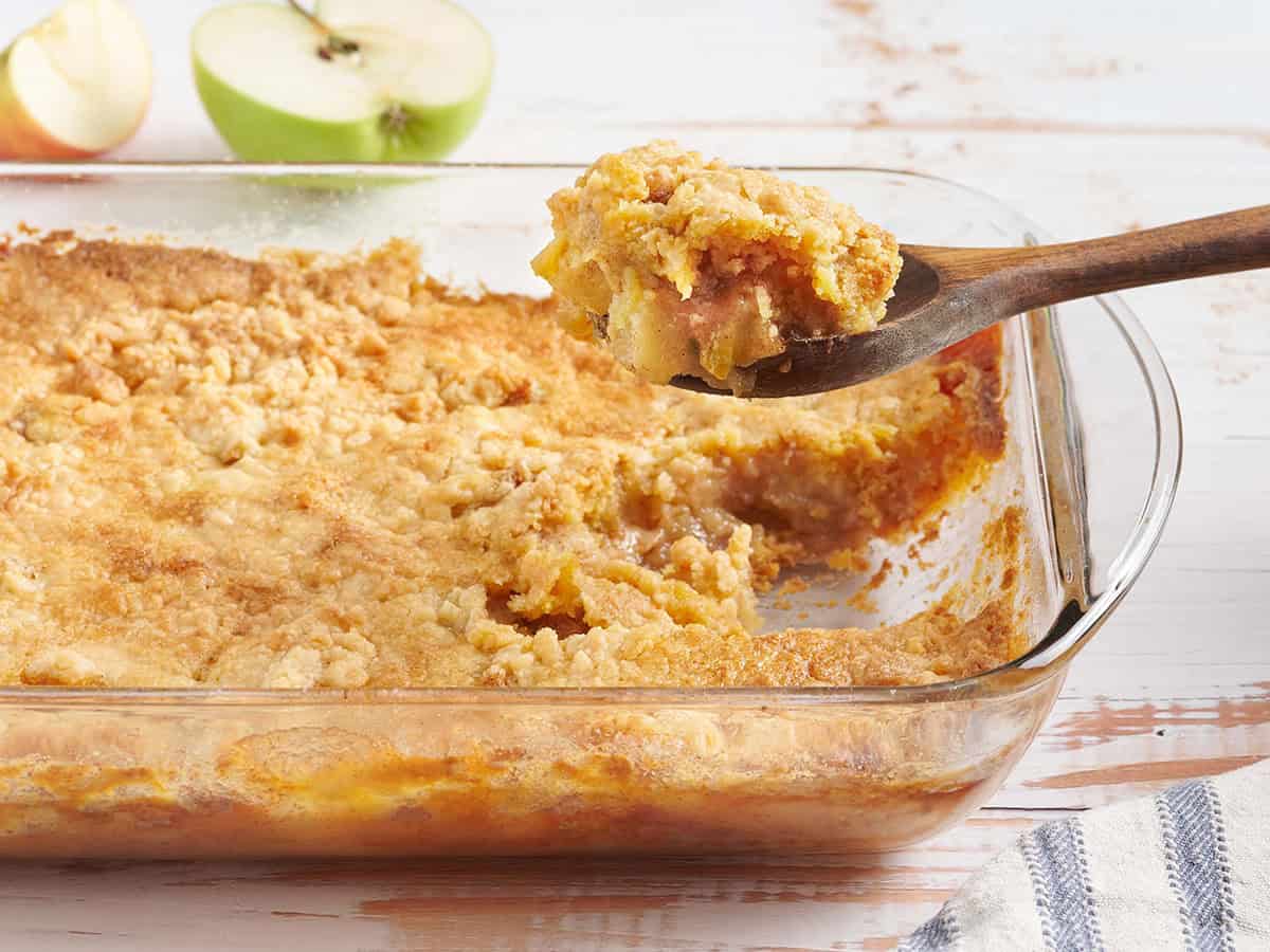 A golden apple dump cake in a glass baking dish with crisp buttery edges and a scoop of vanilla ice cream melting on top, on a warm wood kitchen table