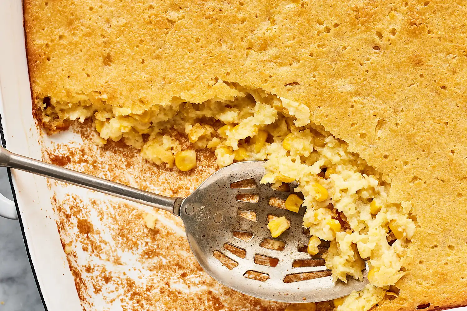 A golden baked Jiffy cornbread casserole in a glass baking dish on a light kitchen counter, with a spoon scooping out a warm, corn-studded portion