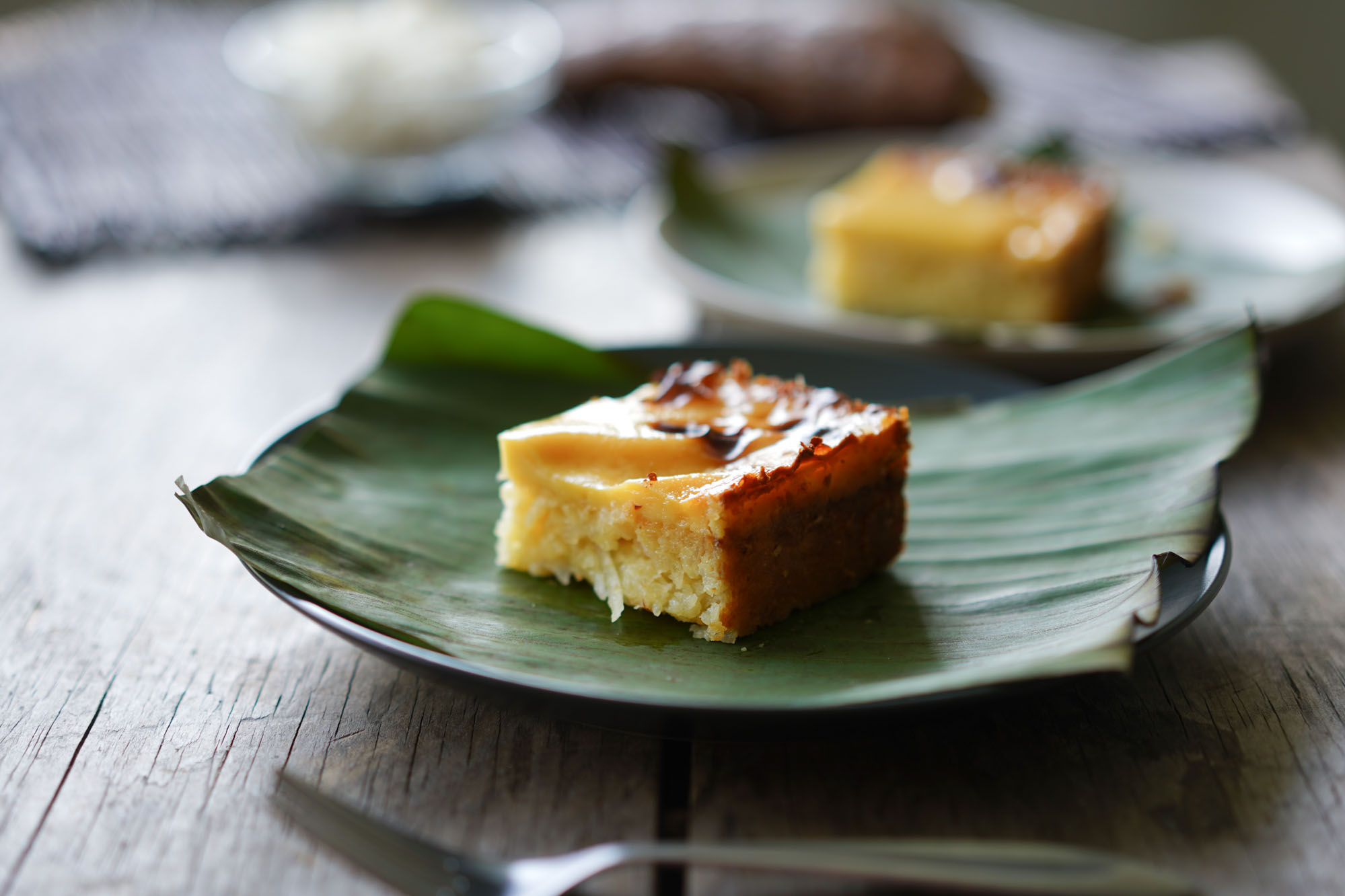 A golden baked cassava cake cut into neat squares on a ceramic plate with a glossy coconut custard top, set on a warm kitchen table with soft natural light