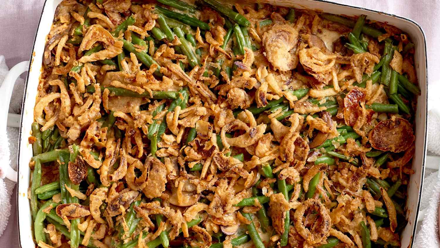 A golden baked green bean casserole in a white ceramic baking dish topped with crisp fried onions on a warm kitchen counter