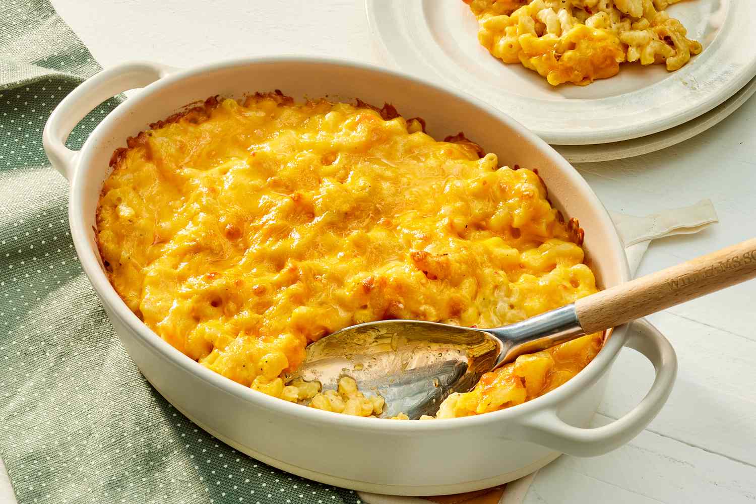 A golden baked macaroni and cheese casserole in a white baking dish on a cozy kitchen table, with crisp browned edges and a spoon scooping out a creamy serving