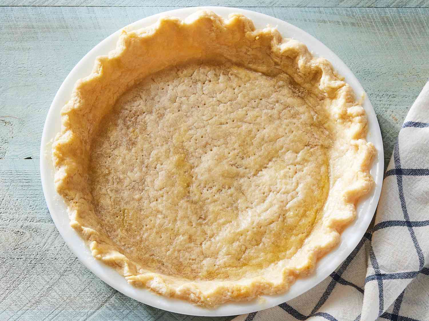 A golden baked pie shell in a glass pie dish on a light kitchen counter with a few scattered flour smudges and a rolling pin nearby