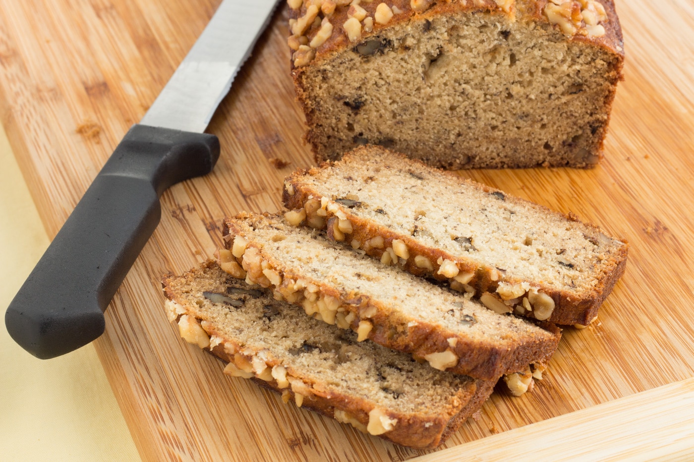 A golden banana nut bread loaf sliced on a wooden cutting board with visible walnut pieces and a few ripe bananas nearby on a bright kitchen counter