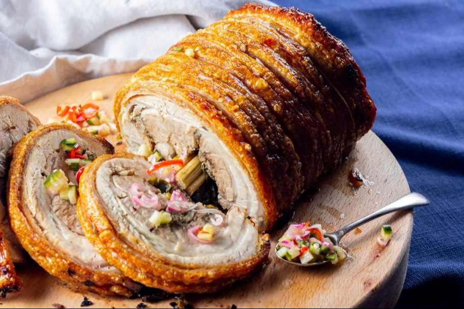A golden, blistered lechon pork belly roast on a cutting board with a small bowl of liver sauce on the side, warm kitchen lighting